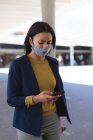 Donna afroamericana che indossa maschera facciale utilizzando smartphone per strada. stile di vita durante il coronavirus covid 19 pandemia. — Foto stock