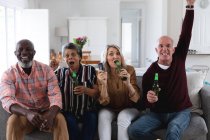 Casais seniores caucasianos e afro-americanos sentados no sofá assistindo ao jogo bebendo cerveja em casa. sênior aposentadoria estilo de vida amigos socialização. — Fotografia de Stock
