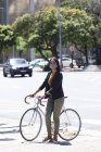 Mujer afroamericana con máscara facial caminando en bicicleta por la calle. estilo de vida durante la pandemia de coronavirus covid 19. - foto de stock