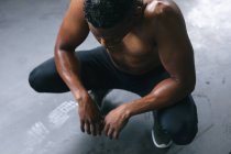 Hombre afroamericano con ropa deportiva en cuclillas descansando después de luchar contra cuerdas en un edificio urbano vacío. aptitud urbana estilo de vida saludable. - foto de stock
