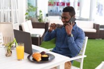 Un Afro-Américain assis dans un café qui parle avec son smartphone et boit du café. homme d'affaires en déplacement dans la ville. — Photo de stock
