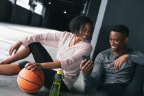 Homem e mulher afro-americanos sentados em prédio urbano vazio e descansando depois de jogar basquete. usando smartphone e rindo. fitness urbano estilo de vida saudável. — Fotografia de Stock