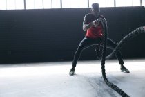 Uomo afroamericano che indossa vestiti sportivi combattendo corde in un edificio urbano vuoto. fitness urbano stile di vita sano. — Foto stock