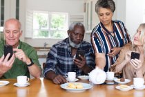 Senior caucasian and african american couples sitting by table drinking tea using smartphone at home. senior retirement lifestyle friends socializing. — Stock Photo