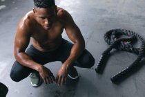 Afroamerikanischer Mann in Sportkleidung hockt nach einem Kampf gegen Seile in einem leeren städtischen Gebäude in der Hocke. urbane Fitness gesunder Lebensstil. — Stockfoto
