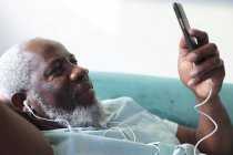Homem americano africano sênior deitado no sofá usando smartphone ouvindo música em fones de ouvido. ficar em casa em auto-isolamento durante o confinamento de quarentena. — Fotografia de Stock