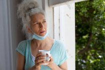 Mujer caucásica mayor de pie junto a la ventana bebiendo una taza de café en casa. permanecer en casa en aislamiento durante el bloqueo de cuarentena. - foto de stock