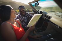 Couple diversifié par une journée ensoleillée assis dans une voiture convertible regardant une carte. road trip estival sur une autoroute de campagne au bord de la côte. — Photo de stock