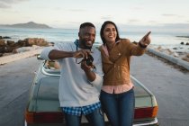 Diverse couple standing by a convertible car and taking photos with camera. Summer road trip on a country highway by the coast. — Stock Photo