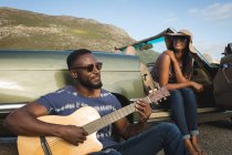 Diverse couple taking roadside break on sunny day beside convertible car the man playing guitar. summer road trip on a country road by the coast. — Stock Photo
