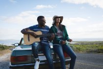 Diverse couple taking roadside break on sunny day beside convertible car the man playing guitar. Summer road trip on a country road by the coast. — Stock Photo