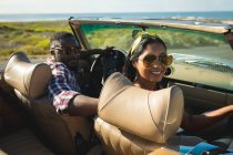 Couple divers conduisant sur une journée ensoleillée en voiture décapotable regardant la caméra et souriant. Road trip estival sur une autoroute de campagne au bord de la côte. — Photo de stock