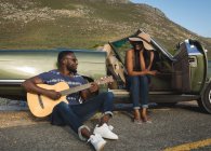 Разнообразная пара отдыхает на обочине дороги в солнечный день рядом с перевернутой машиной, мужчина играет на гитаре. летняя поездка по проселочной дороге на побережье. — стоковое фото
