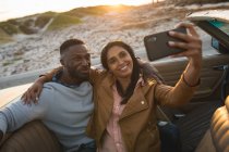 Diverse couple sitting in a convertible car and taking a selfie. Summer road trip on a country highway by the coast. — Stock Photo