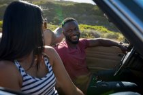 Couple diversifié conduisant sur une journée ensoleillée en voiture décapotable se regardant et souriant. road trip estival sur une autoroute de campagne au bord de la côte. — Photo de stock