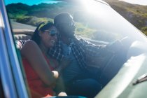 Diverso casal dirigindo no dia ensolarado em carro conversível abraçando e sorrindo. Viagem de estrada de verão em uma estrada rural pela costa. — Fotografia de Stock