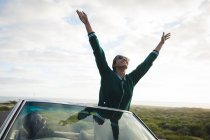 Diverse couple driving on sunny day in convertible car woman is standing and holding her hands up. Summer road trip on a country highway by the coast. — Stock Photo