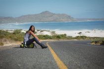 Mixed race woman sitting by the road and hitchhiking. Summer travels on a country highway by the coast. — Stock Photo