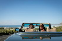 Couple divers conduisant sur une journée ensoleillée en voiture décapotable regardant la caméra et souriant. Road trip estival sur une autoroute de campagne au bord de la côte. — Photo de stock