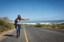 Donna mista che cammina lungo la strada e fa l'autostop. estate viaggia su un'autostrada di paese dalla costa. — Foto stock