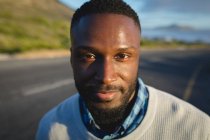 Portrait d'un homme afro-américain debout sur la route regardant la caméra et souriant. été voyage sur une autoroute de campagne par la côte. — Photo de stock