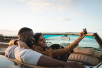 Casal diverso sentado em um carro conversível e tirando uma selfie. Viagem de estrada de verão em uma estrada rural pela costa. — Fotografia de Stock