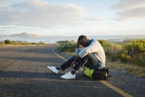 Uomo afroamericano seduto vicino alla strada sul suo zaino. L'estate viaggia su un'autostrada di paese dalla costa. — Foto stock