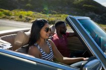 Couple diversifié conduisant sur une journée ensoleillée en voiture convertible parler et sourire. Road trip estival sur une autoroute de campagne au bord de la côte. — Photo de stock