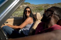 Diversa pareja conduciendo en un día soleado en un coche descapotable mirándose y sonriendo. viaje de verano por carretera en una carretera rural junto a la costa. - foto de stock