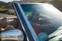Mixed race woman driving on sunny day in convertible car holding driving wheel. Summer road trip on a country highway by the coast. — Stock Photo