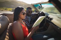 Diverse couple on sunny day sitting in convertible car looking at a map. summer road trip on a country highway by the coast. — Stock Photo