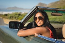 Смешанные гонки женщина за рулем в солнечный день в конвертируемом автомобиле, держа за руль и улыбаясь. Летняя поездка по загородному шоссе у побережья. — стоковое фото