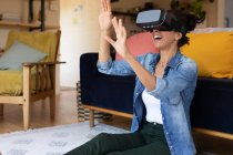 Mujer caucásica sonriendo usando auriculares vr sentados en el suelo en casa. Permanecer en casa en aislamiento durante el bloqueo de cuarentena. - foto de stock