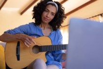 Donna caucasica che suona la chitarra usando il computer portatile seduto sul divano a casa. stare a casa in isolamento durante l'isolamento in quarantena. — Foto stock