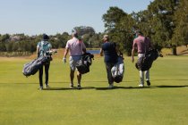 Cuatro hombres y mujeres mayores caucásicos con máscaras faciales caminando por el campo de golf sosteniendo bolsas de golf. deporte de golf hobby, estilo de vida de jubilación saludable durante coronavirus covid 19 pandemia. - foto de stock