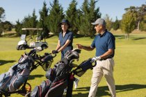 Homem e mulher idosos caucasianos que atravessam o campo de golfe com sacos de golfe. passatempo de esportes de golfe, estilo de vida de aposentadoria saudável — Fotografia de Stock