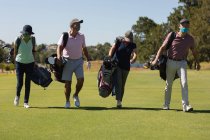 Четверо белых мужчин и женщин в масках для лица ходят по полю для гольфа с сумками для гольфа. гольф спортивное хобби, здоровый пенсионный образ жизни во время коронавируса ковид 19 пандемии. — стоковое фото