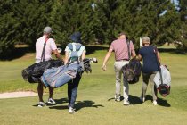 Cuatro hombres y mujeres mayores caucásicos caminando por el campo de golf sosteniendo bolsas de golf. deporte de golf hobby, estilo de vida de jubilación saludable durante coronavirus covid 19 pandemia. - foto de stock