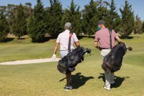 Dois homens caucasianos com máscaras faciais a atravessar o campo de golfe com sacos de golfe. passatempo esportivo de golfe, estilo de vida de aposentadoria saudável durante coronavírus covid 19 pandemia. — Fotografia de Stock