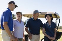 Четыре белых старших мужчины и женщины стоят у гольф-багги, держа клюшки для гольфа и разговаривая. Спортивное увлечение гольфом, здоровый пенсионный образ жизни — стоковое фото