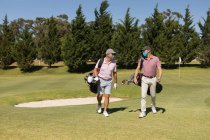 Двоє кавказьких літніх чоловіків у масках для обличчя, що ходять полем для гольфу, тримають сумки для гольфу. Гольф спортивне хобі, здоровий спосіб життя на пенсії під час пандемії коронавірусу 19 . — стокове фото