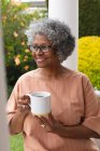 Старшая африканская женщина держит чашку кофе, улыбаясь, сидя на крыльце дома. оставаться в изоляции в карантинной изоляции — стоковое фото