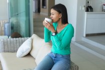 Femme afro-américaine buvant du café assis sur le canapé à la maison. rester à la maison en isolement personnel en quarantaine — Photo de stock