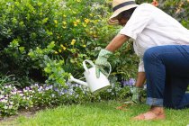 Mujer mayor afroamericana con guantes de jardinería sonriendo mientras regaba plantas en el jardín. permaneciendo en aislamiento en cuarentena - foto de stock