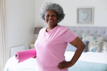 Ritratto di donna anziana afroamericana sorridente mentre tiene in mano il tappetino yoga a casa. stare a casa in isolamento personale in quarantena — Foto stock