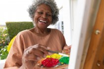 Mujer mayor afroamericana sonriendo mientras pinta sobre lienzo de pie en el porche de la casa. permaneciendo en aislamiento en cuarentena - foto de stock