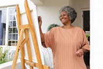 Eine afroamerikanische Seniorin lächelt, während sie auf der Veranda des Hauses auf Leinwand malt. Isolation in Quarantäne — Stockfoto