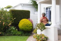 Mujer mayor afroamericana reflexiva sosteniendo una taza de café mientras está sentada en el porche de la casa. permaneciendo en aislamiento en cuarentena - foto de stock
