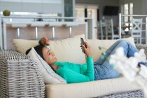 Mujer afroamericana usando teléfono inteligente acostado en el sofá en casa. permaneciendo en casa en aislamiento en cuarentena - foto de stock
