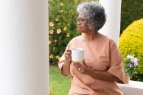 Nachdenkliche afrikanisch-amerikanische Seniorin mit Kaffeetasse auf der Veranda des Hauses sitzend. Isolation in Quarantäne — Stockfoto